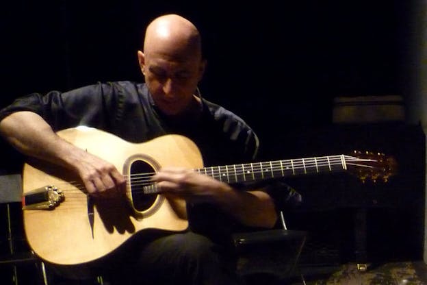 Elliot Sharp is in the center, playing guitar. He wears a dark shirt and sits in front of a black background.