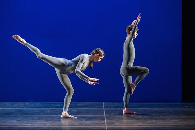 Two performers in gray unitards strike poses against a matte blue background: one faces sideways and balances on one bent knee, raising both arms in a v-shape above their head; the other bends on one knee towards the ground, palms facing the floor, their right leg extended in arabesque with a pointed foot.