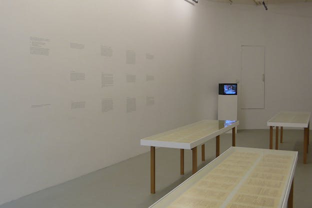 Installation view of three glass-framed tables displaying yellow text-filled papers beside a white wall covered in blocks of indiscernable black text and in the back, a small television displaying a person's face with indiscernable white subtitles. 