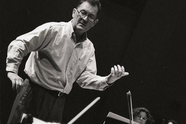 Black and white photograph of a conductor standing and motioning with his hands to musicians sitting down with their instruments behind music sheets.