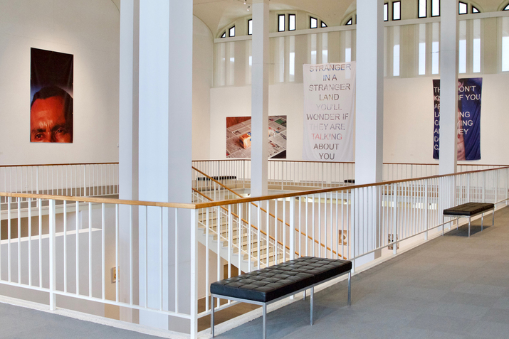 An exhibition balcony with various images hanging from the walls. The image central to the viewer near the stairs reads 