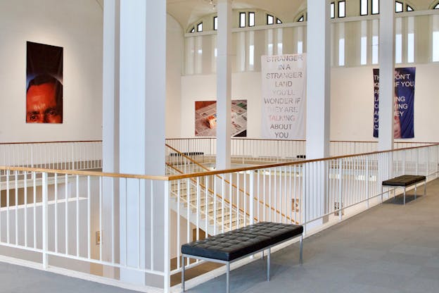 An exhibition balcony with various images hanging from the walls. The image central to the viewer near the stairs reads 
