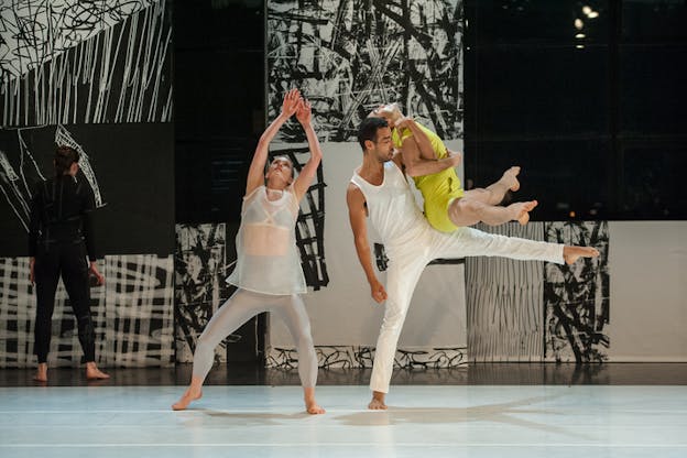 On a bright stage set with a backdrop of black and white abstract paintings, one performer balances another performer on their extended right leg, one performer bends their knees and looks upwards at their raised arms, and one performer stands upstage, facing the back wall.