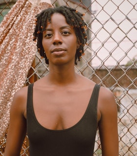 A portrait of Marguerite Hemmings wearing a blank tank top and  translucent orange beads in their hair. They are standing in front of a wire fence with a bit of rose gold sequined fabric hanging off its edge. They face the camera directly and smile slightly.