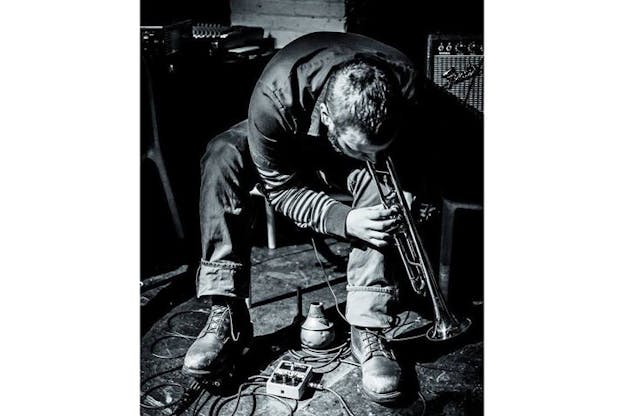 Black and white photograph of Wooley sitting and bending over, playing the trumpet tilted towards the floor. 