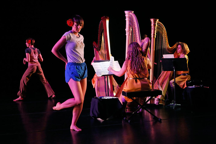 Three seated figures play harps as two performers, one in the front and one in the back, with red flowers in their hair stand in different positions.  