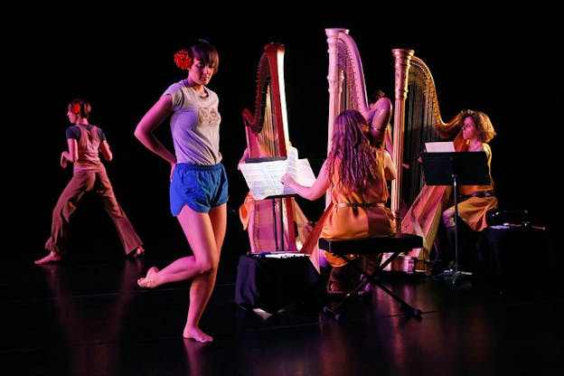 Three seated figures play harps as two performers, one in the front and one in the back, with red flowers in their hair stand in different positions.  