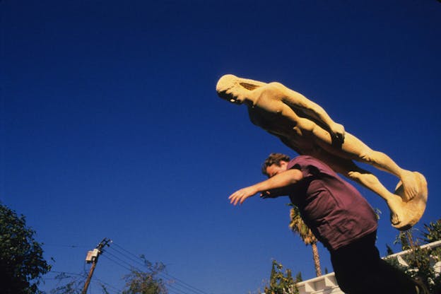 A photogrtaph taken at a low angle of Martin Kersels leaning forward with a statue like a greek kouros on top of his back. As a result of the angle, all that is visible of the background are telephone wires, the top of a white building and the blue sky. Kersels wears a burgundy top and dark pants. 