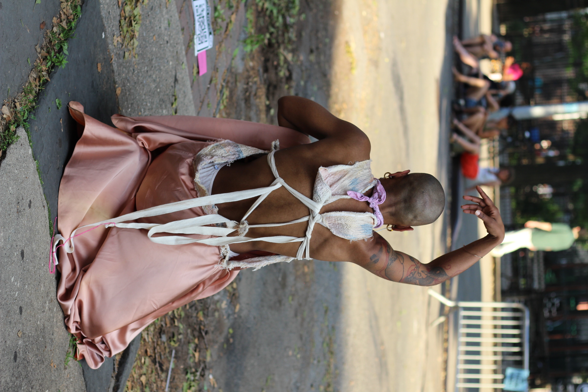 Jasmine Hearn kneels outdoors on a cement tile next to a street, their back to the camera with one hand hanging above their head. They wear a pink skirt and a white top tied loosely behind their back.