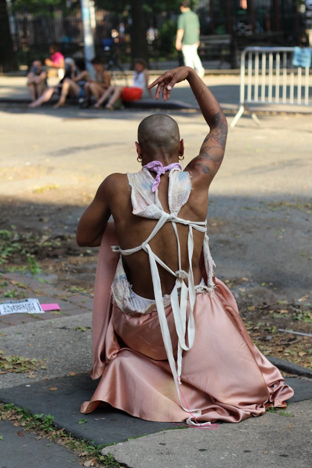 Jasmine Hearn kneels outdoors on a cement tile next to a street, their back to the camera with one hand hanging above their head. They wear a pink skirt and a white top tied loosely behind their back.