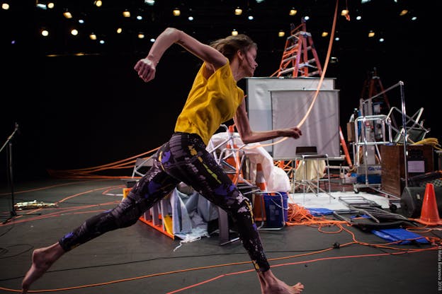 Joanna Kotze performs on a brightly lit stage filled with equipment including projection screens, ladders, clothing racks, orange cones, and electrical cords among other things. She is centered in the frame in profile view as she runs to the right. Her right leg strides forward as her left trails behind her, her arms are bent at the elbows, her right swings behind her as her left reaches forward. She is barefoot, wearing a sleeveless yellow top and black and purple abstract patterned leggings. 