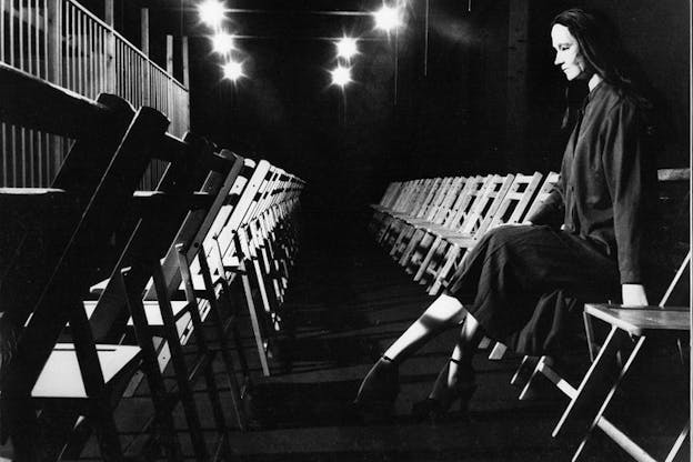 A person dressed in a gray suit dances and twists their body in the center of a black stage with gray floors while a person standing in the back left smiles and watches. 