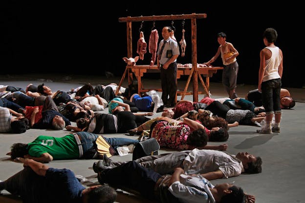 A performance still of people laying on the ground, in various street clothes besides a wooden frame with bits of raw meat hanging from it. Three performers are standing. One of them is front of the frame, and is dressed in a blue short sleeve button up shirt and a tie. Behind the wooden frame, a downtrodden looking performer wearing grey pants and a smudged white t-shirt leans on the wooden frame. A third performer stands facing the frame, wearing street clothes and a plastic bag wrapped around his left sneaker. 