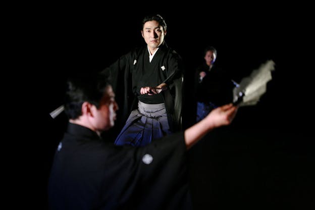 A performance still of a three performers in a darkened room, wearing kimonos. Each performer holds a fan in their outstretched arm and looks forward intently. Two of the performers look directly at the audience and the other is turned sideways. The performer in the foreground and background are blurred.  