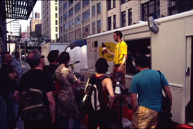 A performer stands in from of a white truck parked on a city street. The truck has a yellow sticker on it which reads 