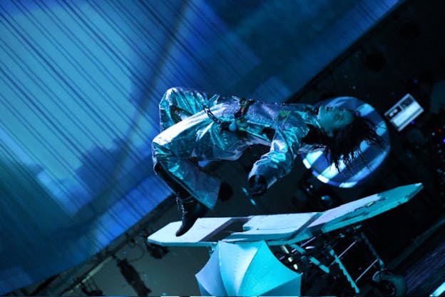 Sideways angle of a performer in a shiny silver suit appearing to levitate above a hospital bed in a blue-hued room.