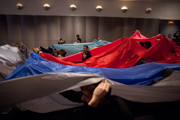People in a large gray room hold sweeping red, blue, and gray tarps over their heads.