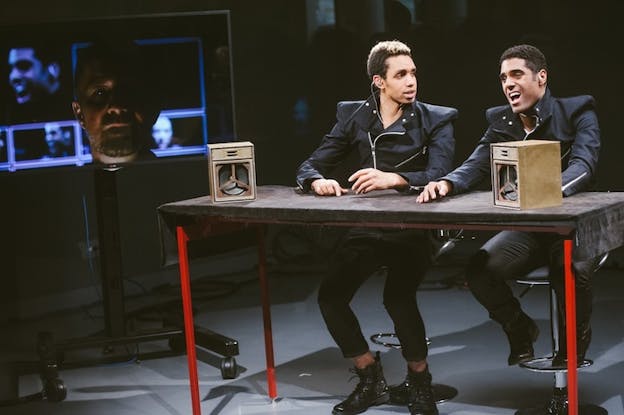 Two people sit next to each other on a desk with red legs. In front of each one stands a wooden box, with a mechanism inside. Next to the on the TV the face of a person, mostly shadowed.