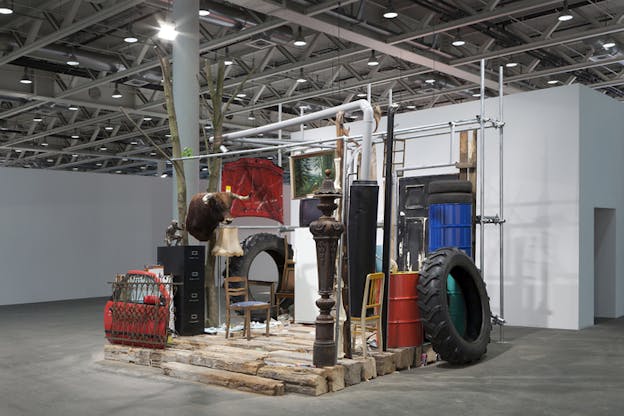 Exhibition containing tires, paintings, chairs and a car door situated on top of wooden logs.