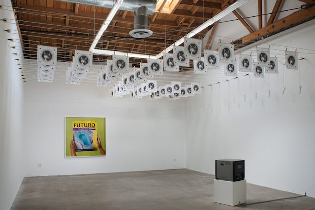 Gallery room with images of a cat's head hanging from the ceiling. Underneath it on a white square a gray electronic box. In the wall facing the viewer an image with hands holding a blue item on a grenn background and the word 
