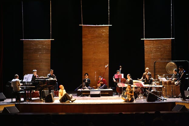 An orchestra and some performers on the floor, perform on a stage with a black background adorned with three aligned brown wooden parchments hanging from the ceiling.