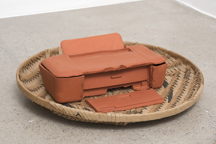 A cirlce weaved basket with brown,black and blue colors holds inside it an orange clay printer.