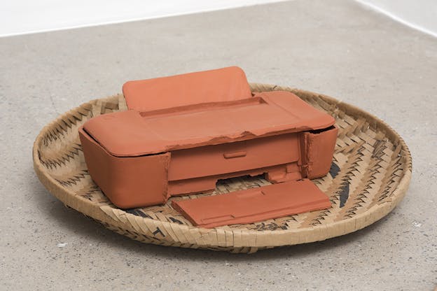 A cirlce weaved basket with brown,black and blue colors holds inside it an orange clay printer.
