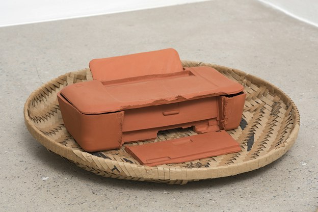 A cirlce weaved basket with brown,black and blue colors holds inside it an orange clay printer.