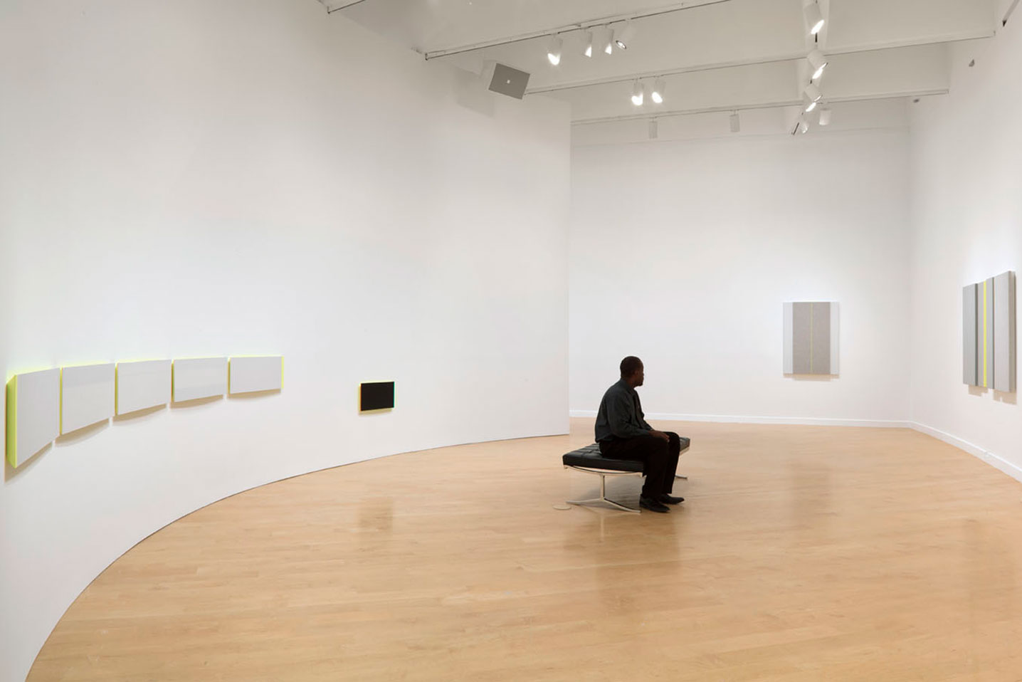A person in dark clothing with their head facing away from the viewer sits on a gallery room bench. Surrounding them painting of white-gray color with lines of neon yellow in different shapes.