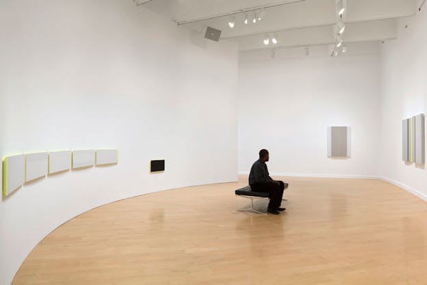 A person in dark clothing with their head facing away from the viewer sits on a gallery room bench. Surrounding them painting of white-gray color with lines of neon yellow in different shapes.