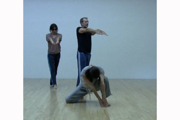 Two performers in the back cross their feet and outstretch their arms while a third performer kneels and bends their head downwards, their fingers touching the ground.