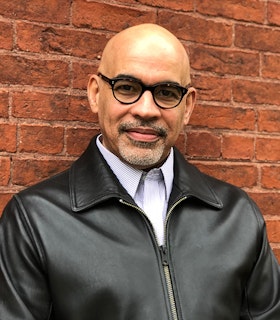 A portrait of Willie Perdomo in front of a red brick wall. He is clad in a black leather jacket with black-framed glasses adorning his eyes.
