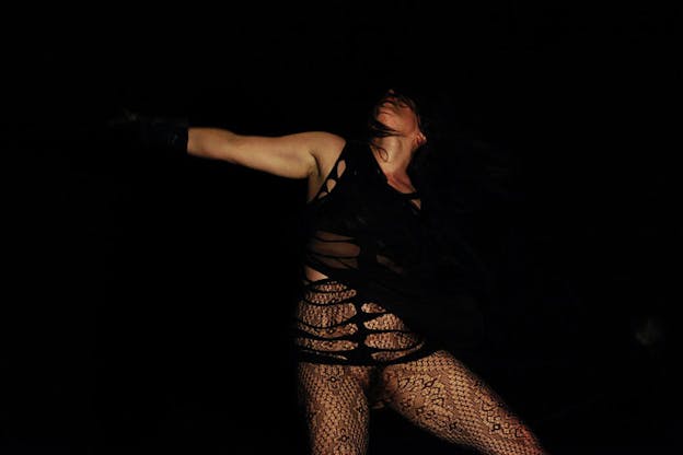 Performer whipping their head back, their hair covering their eyes, outstretching one arm, dressed in black lace clothing that blends into pitch black space. 