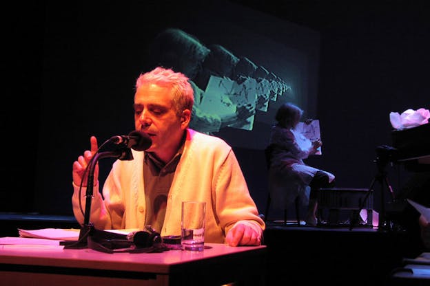 One performer sits at a desk and speaks into a microphone. They have their eyes closed and are raising their right hand and pointing their right index finger. They are iluminated in warm orange and pink light. Behind them, a person holds a small drawing on paper. In a projection behind them, their image is copied and shown many times side by side. This scene is bathed in cool-toned blue and purple light. 