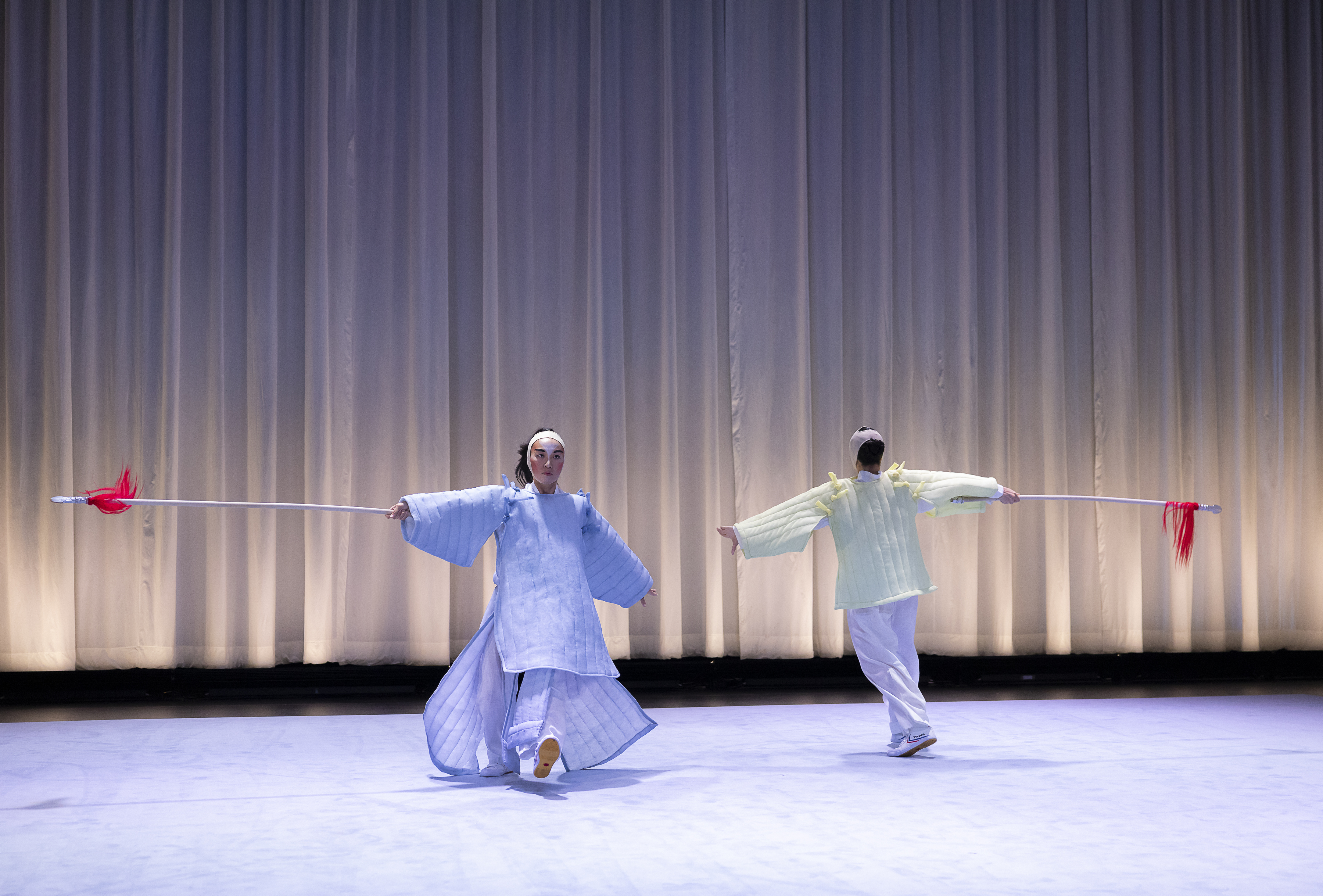 Lai Yi Ohlsen and Pareena Lim performing, moving in a circle.  They are both wearing costumes inspired by Eastern Asian performance  traditions. Lai Yi Ohlsen is facing the audience is in light purple dress, while  Pareena Lim, facing back stage, is in light yellow. Both of their hands are in  the air to their sides, and they each have one foot in front of the other.  Behind the duo is a white curtain that is just a bit short of reaching the  ground, the pitch-black of back stage is visible from below.  The floor is light purple, matching Lai’s dress.