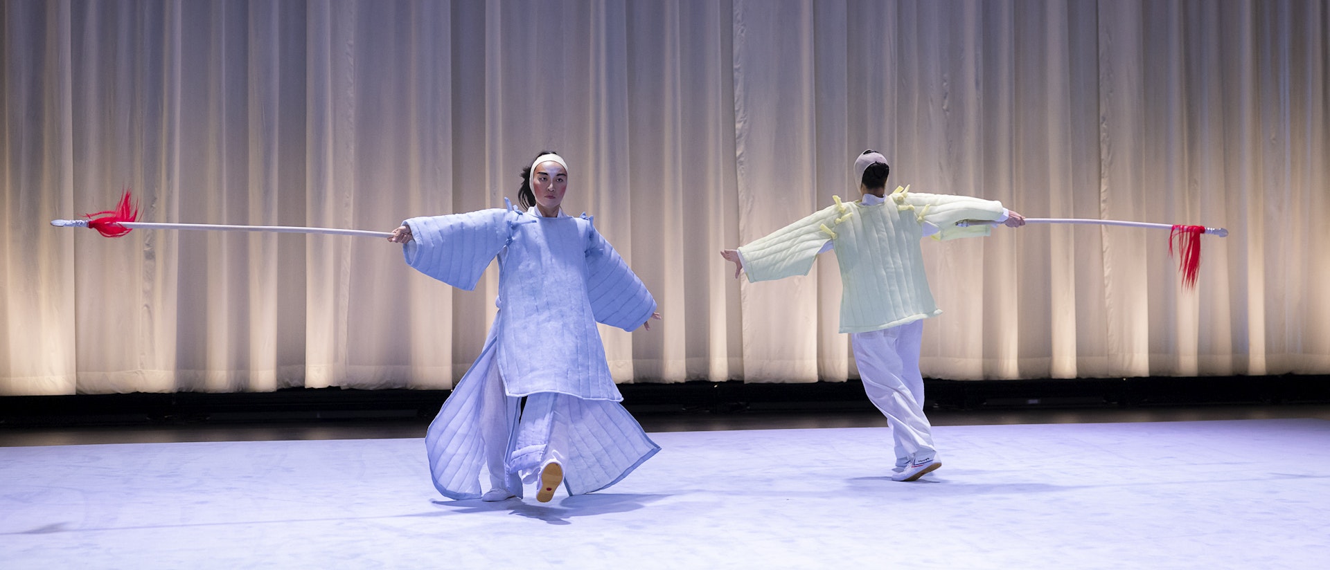 Lai Yi Ohlsen and Pareena Lim performing, moving in a circle.  They are both wearing costumes inspired by Eastern Asian performance  traditions. Lai Yi Ohlsen is facing the audience is in light purple dress, while  Pareena Lim, facing back stage, is in light yellow. Both of their hands are in  the air to their sides, and they each have one foot in front of the other.  Behind the duo is a white curtain that is just a bit short of reaching the  ground, the pitch-black of back stage is visible from below.  The floor is light purple, matching Lai’s dress.