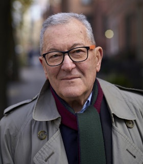 A portrait of Petr Kotik in front of a blurred city street. He wears a grey trench coat, a navy blue sweater, a red and green scarf, and black and orange glasses.