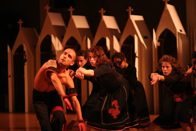 One performer wearing black pants and red and black braided arm and wrist bands leans to the right in a half-kneeling position. Behind them, multiple performers wearing red and black sleeve dresses squat with extended arms and stare directly to the front. Some of the dresses are printed with a red rooster motif. Horizontally lined up against a black background, six beige color arch structures with triangular roofs each has a small cross placed at the top end.