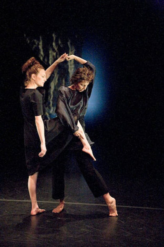 One performer wearing a black midi t-shirt dress raises their right thigh up in a 90-degree angle while looking to their right. A second performer wearing a black long-sleeve shirt and pants grabs the right calf of the other performer while looking down. The two holds their left hands together above their heads. The performance is staged on a black floor against a black backdrop.