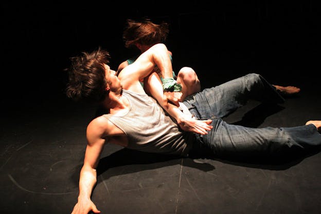 Two performers are on a black floor. One performer leans on their right arm and clutches the other performer's leg with their left arm. They look behind them at the other performer who is crouched on the floor. 