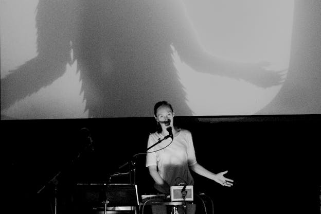 Black and white photograph of a person behind a booth speaking on a microphone with their arms slightly raised to their sides. Behind them the projection of a silhouette with fur doing the same motion.