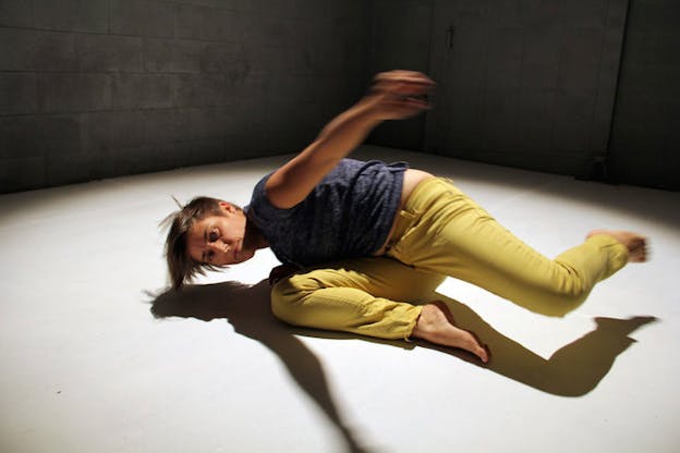 Performer in a gray shirt and yellow pants falls to the ground, their right arm and leg outstretched and blurred in motion. 