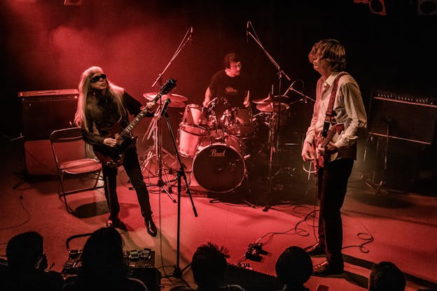 Three musicians play drums and guitar, bathed in red light. Several microphones, two amplifiers, and a folding chair are onstage with them.