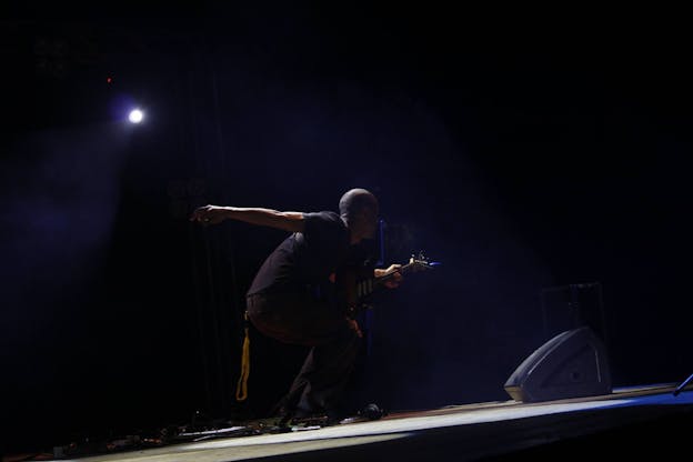 Morgan Craft plays the guitar on a dark stage illuminated by a spotlight, body facing towards the right, away from the camera. He is leaning forward with bent knees, his right arm is extended behind him while his left hand holds his guitar. He is wearing a dark t-shirt and black jeans, an orange lanyard hangs from his back pocket.
