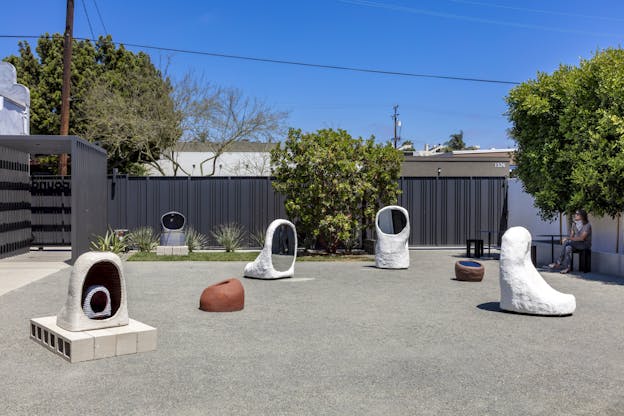 An installation view of seven of Anna Sew Hoy’s sculptures installed in an outdoor gravel-lined sculpture garden. Each sculpture is an organic shape with circular mirrored elements.