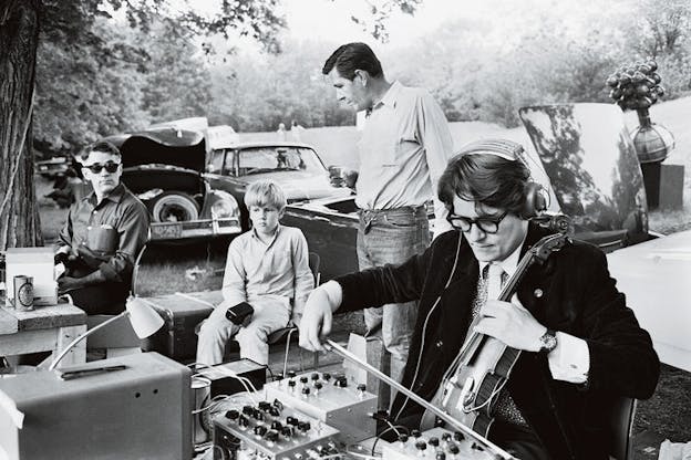 Several performers sit in an open natural space with many trees in the far background. In the foreground, Gordon Mumma plays violin and wears thick black glasses and a suit. He sits in front of a sound box. To the left of him, John Cage stands and looks over a Mark Nelson who sits and David Tudor who looks on while wearing sunglasses. 