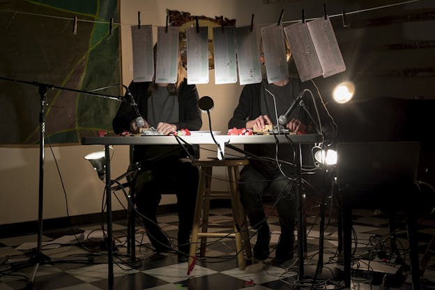 Two figures clad in black facing the viewer sit behind a table with things in their hands and microphones on top of them. Longs papers hangs from a rope covering their faces from view. A lamp is lit behind them, while a computer on a stand and black tangled cables sit on the floor in front of them.