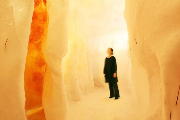 A figure dressed in black baggy clothes stand inside a fot yellow and orange space with smooth rocks compromising the walls. The person's face is blurred and right is softly blurred.