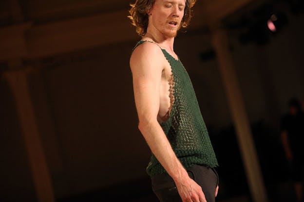 A performer wearing a delicate green tank top patterned with small green dots stands with their arms at their side and looks down with an exhausted expression on their face. The background is dark and blurred.