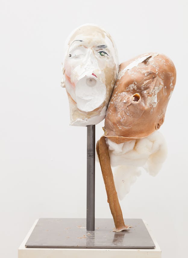 A plaster head sits atop a metal rod. The mouth and most of the left side of the head is rubbed off but a single green eye is visible on the right. Leaning against this head is another head made of peach colored latex, with fewer facial features. Fluffy material comes out of the neck of the head and the latex rod that this head is balanced on is bent. 